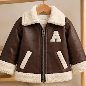 Könnte beinhalten: Eine dunkelbraune Jacke mit einem cremefarbenen Kragen, Ärmelbündchen und Saum aus Kunstfell. Die Jacke verfügt über einen zentralen Metallreißverschluss, zwei schräge Vordertaschen mit cremefarbenem Besatz und einen großen, weißen Buchstaben A.