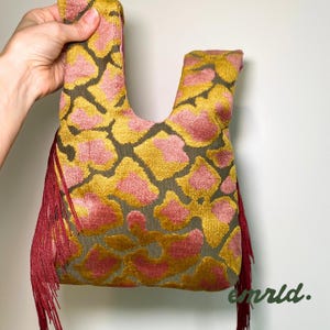 Puede incluir: Un bolso de tela con un patrón texturizado similar al de una jirafa en tonos amarillo mostaza y rosa empolvado sobre un fondo oscuro. El bolso tiene un asa integrada y largos flecos de color rojo oscuro en los laterales.
