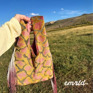 May include: A soft, textured handbag featuring a giraffe-like pattern in shades of mustard yellow and dusty pink, with long, gradient-dyed fringe detailing on the sides and a solid pink interior lining.