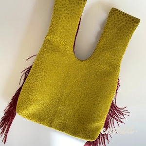 May include: A mustard yellow handbag with a textured, dimpled surface and a Japanese-style knot handle. The bag features long, deep red fringe trim hanging from both sides, creating a distinct contrast against the yellow fabric.