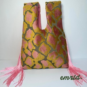 Puede incluir: Un bolso tote de tela texturizada similar al terciopelo con un estampado animal abstracto en rosa y amarillo mostaza. El bolso tiene asas integradas y largos flecos rosas fluidos en ambos lados. El texto emrld. es visible.