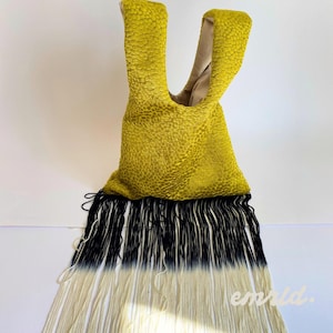 Puede incluir: Un bolso de tela texturizada color chartreuse con asa tipo nudo y forro interior beige. La base del bolso presenta largos flecos colgantes que pasan de negro en la parte superior a un color blanco roto en los extremos inferiores.