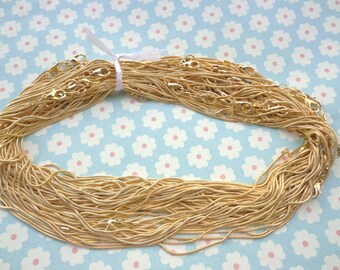 May include: A large pile of gold snake chain necklaces with clasps. The chains are arranged in a circle and are tied together with a white ribbon.