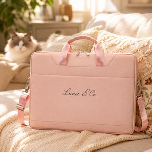 Peut inclure: Un sac pour ordinateur portable rectangulaire rose clair doté de deux poignées supérieures et d'une bandoulière amovible. L'avant du sac affiche le texte Luna & Co. en police cursive noire. Le sac possède une fermeture éclair avec des détails argentés.