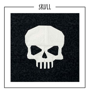 May include: A square fabric quilt block featuring a white skull with a subtle dot pattern centered on a black background with a tonal swirling floral print. The word SKULL is printed in black capital letters above the square fabric design.