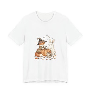 May include: A white short-sleeve t-shirt featuring a central graphic illustration of a fox wearing a witch hat, a rabbit dressed as a ghost, and a hedgehog wrapped like a mummy, all gathered around a large orange pumpkin with autumn leaves.