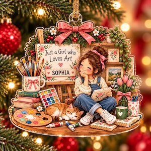 May include: A clear acrylic Christmas ornament featuring a girl with a pink hair bow sitting by an easel. The sign reads Just a Girl who Loves Art and SOPHIA. Includes a sleeping orange cat, paintbrushes, a palette, books, and flowers.