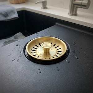 Könnte beinhalten: Ein kreisförmiges Küchenspülen-Sieb aus Messing mit einem zentralen Knopf und einer Reihe geschwungener Abflusslöcher. Das goldfarbene Metallsieb ist in ein mattes, schwarzes Spülbecken eingelassen, mit kleinen Wassertropfen auf der Oberfläche.