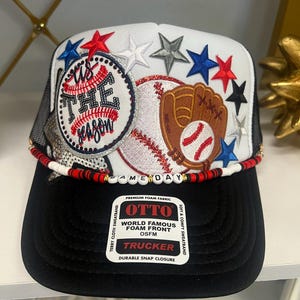 May include: A white and black trucker hat featuring a baseball graphic with the text 'tis THE season', a brown baseball glove, and various red, white, blue, and silver star patches. A beaded cord with the text 'GAME DAY' sits above the black brim.
