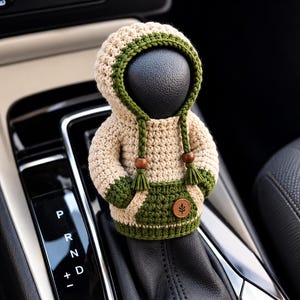 Peut inclure: Une housse de levier de vitesse en crochet beige et vert foncé. Le vêtement présente une capuche avec des cordons tressés terminés par des perles en bois et des pompons, une poche kangourou et une petite étiquette ronde marron avec une feuille.