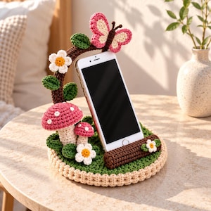 Könnte beinhalten: Ein Smartphone-Ständer aus Häkelarbeit mit einer runden grünen Basis und einer braunen Stütze in Holzoptik. Er ist verziert mit zwei rosa-weiß gepunkteten Pilzen, kleinen weißen Blumen mit gelber Mitte, grünen Blättern und einem rosa Schmetterling.