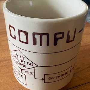 May include: A cream-coloured ceramic mug featuring dark brown text and a flowchart graphic. The text reads COMPU- at the top, with a decision diamond asking IS IT TIME TO GO HOME, leading to a box that says GO HOME and a box for COFFEE BREAK.