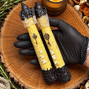 May include: Two cylindrical yellow candles featuring a black wax drip design at the top and bottom. The top sections are wrapped in a textured, translucent material, while the centre of each candle is adorned with a small bronze-coloured key charm.