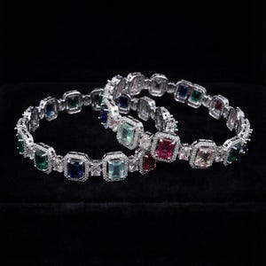 May include: Two silver-toned bangles featuring a series of rectangular-cut multi-colored gemstones, including emerald green, sapphire blue, ruby red, and pale pink, each surrounded by a border of small, clear, sparkling white cubic zirconia stones.