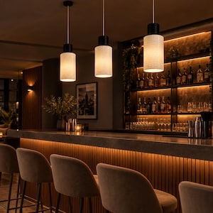 Pode incluir: Três candeeiros suspensos cilíndricos com abajures de vidro branco texturizado e ferragens pretas suspensos sobre um balcão de bar. As luzes emitem um brilho quente, iluminando a área do bar com bancos estofados e prateleiras com garrafas.