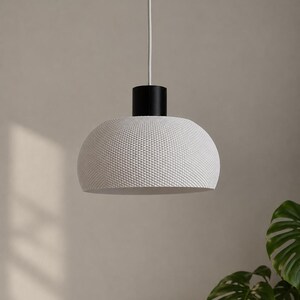 Pode incluir: Uma luminária pendente em forma de cúpula com uma cúpula branca texturizada apresentando um padrão de grade geométrica fina. A cúpula está fixada a um suporte cilíndrico preto e suspensa por um fio branco fino, com um visual limpo e simples.