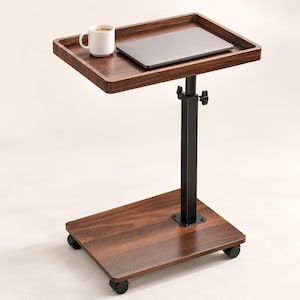 May include: A mobile side table with a dark wood grain finish, featuring a rectangular top and base connected by a black adjustable metal post. The table includes four small caster wheels, a white ceramic mug, and a closed grey laptop on top.