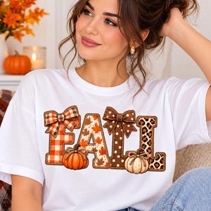 Peut inclure: Un t-shirt blanc à manches courtes avec le mot FALL écrit en grandes lettres. Le F a un motif à carreaux rouges avec un nœud, le A des feuilles d'automne, le L un motif à pois marron avec un nœud, et le second L un imprimé léopard.
