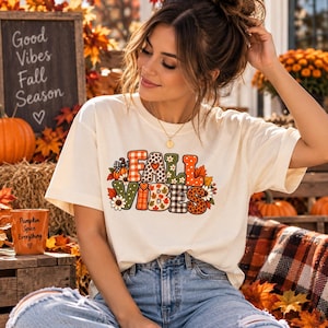 Peut inclure: Un t-shirt à manches courtes de couleur crème avec le texte "FALL VIBES" en grandes lettres. Chaque lettre est remplie de motifs variés, notamment des carreaux orange, des imprimés léopard, des pois, des feuilles d'automne, des citrouilles et des fleurs.