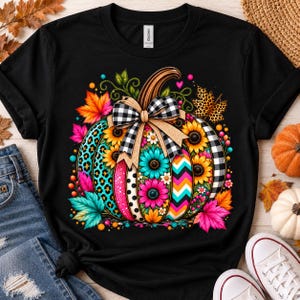 May include: A black short-sleeve t-shirt featuring a large graphic of a pumpkin decorated with sunflowers, leopard print, polka dots, and chevron patterns. A black and white buffalo plaid bow sits on top, surrounded by colourful autumn leaves.