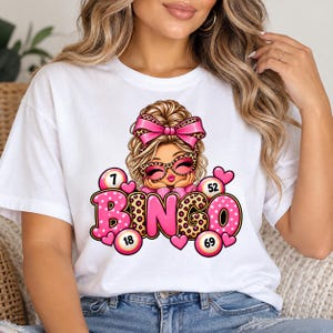 Peut inclure: Un t-shirt blanc avec un motif représentant un personnage de dessin animé aux cheveux blonds en chignon, portant un nœud et des lunettes à motif léopard rose. Le design inclut le mot BINGO en lettres roses et léopard, entouré de boules numérotées.
