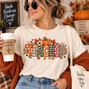 Peut inclure: Un t-shirt crème arborant le mot TEACHER en grandes lettres à motifs, entouré de feuilles d'automne, de fleurs et d'une citrouille. Le t-shirt est porté avec un cardigan rouille, et un gobelet blanc avec le texte Fueled by Students & Coffee est tenu à proximité.