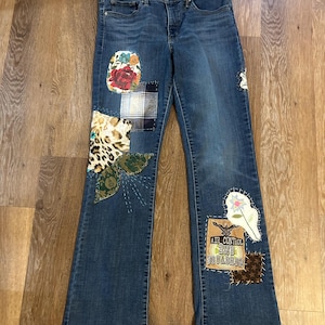 May include: Blue denim jeans featuring a variety of fabric patches sewn onto the legs. The patches include floral, plaid, leopard print, and solid green patterns, along with a rectangular patch displaying the text AIR CONTROL 2ND SQUADRON.