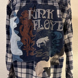 May include: A long-sleeve button-down shirt in a navy blue and white plaid pattern. The back features a large dark blue fabric patch with the text "PINK FLOYD" in a stylized font, a brown illustration of a figure, and a white graphic of a pig.