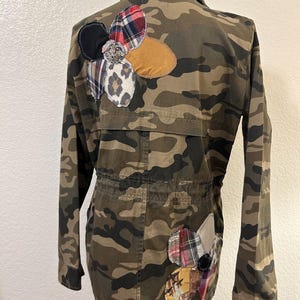May include: A camouflage print utility jacket featuring long sleeves, a collar, and a drawstring waist. The back of the jacket is decorated with two large floral-shaped appliqués made from various fabric patches, including plaid, leopard print, and solid tan.