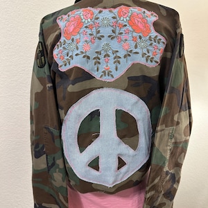 May include: A camouflage print jacket featuring a large light blue fabric patch with pink and green floral embroidery on the upper back and a circular light blue fabric patch with a peace sign symbol on the lower back area.