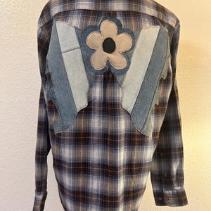 May include: A long-sleeve button-down shirt in a brown, blue, and white plaid pattern. The back features a large, light-colored fabric flower appliqué with a dark center, layered over several vertical strips of light and medium blue denim fabric.