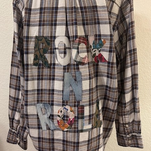 May include: A long-sleeve button-down shirt featuring a brown, white, and blue plaid pattern. The back of the shirt displays the text ROCK N ROLL in large, fabric-appliqued letters, each crafted from various patterned and solid-colored textiles.