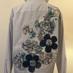May include: A long-sleeved button-down shirt with a light blue and white vertical stripe pattern. The back features a ruffled collar and several floral fabric patches in muted tones, including two dark navy blue denim flower appliqués.