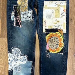 May include: Dark blue denim jeans featuring various fabric patches and visible white hand-stitched embroidery. The patches include floral patterns, a sunflower design, and a rectangular piece with text that reads Love is patient, love is kind.