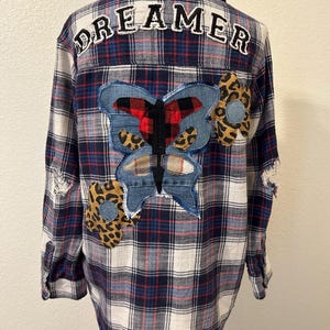 May include: A long-sleeve button-down shirt in a blue, white, and red plaid pattern. The back features the word DREAMER in white letters, a large central butterfly patch made of denim and plaid fabric, and two leopard print flower patches.