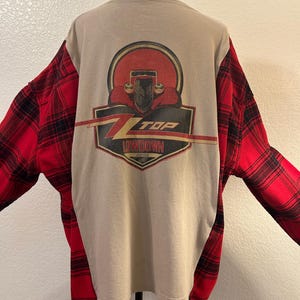 May include: A beige t-shirt with red and black plaid flannel sleeves and side panels. The back features a graphic of a vintage car front with the text Z TOP LOWDOWN in a red and black shield design, centered on the beige fabric panel.