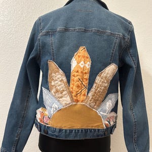 May include: A blue denim jacket featuring a back panel decorated with a sunburst-shaped appliqué made from various fabric scraps in shades of orange, tan, and floral patterns, all secured with visible zigzag stitching along the edges.