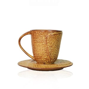 Può includere: Un set di tazza e piattino in ceramica caratterizzato da un motivo testurizzato a tratteggio incrociato nei toni del marrone dorato e del giallo. La tazza presenta un manico curvo e un corpo leggermente affusolato, con un piattino abbinato.