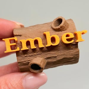 May include: A brown, cylindrical, log-shaped object with a textured bark surface and two small circular protrusions. The word Ember is printed in bright orange, raised, three-dimensional capital letters across the front of the log.