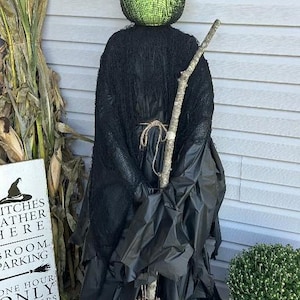 May include: A standing witch decoration featuring a bright green textured head, a tall black pointed hat, and a black draped garment. The figure holds a rustic wooden broom with a natural twig base and is accented with a simple twine tie at the waist.