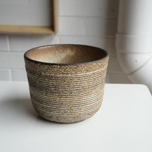 May include: A ceramic bowl with a textured, ribbed exterior featuring horizontal grooves. The surface has a mottled brown and beige finish with a darker, earthy rim. The interior is smooth and light-coloured, contrasting with the rough, tactile outer texture of the vessel.