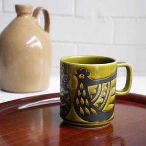 May include: Green ceramic mug featuring a black and gold chicken design. The mug has a handle and is on a wooden tray. A brown jug is in the background. A unique piece of kitchenware.