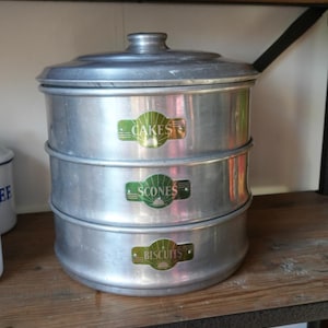 May include: A three-tiered, vintage aluminium cake carrier with a lid. Each tier is labelled with a green and gold sticker: "Cakes," "Scones," and "Biscuits." The carrier sits on a wooden shelf.