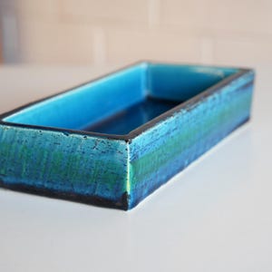 May include: A rectangular ceramic tray featuring a glossy glaze with horizontal bands of deep blue, vibrant turquoise, and emerald green. The interior is a solid, smooth turquoise, while the exterior sides display a textured, crackled finish.