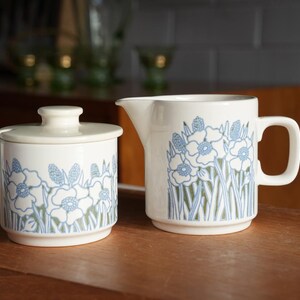 May include: A white ceramic sugar bowl with a lid and a matching creamer jug, both decorated with a repeating blue and green floral pattern featuring stylised flowers and tall grass blades on a smooth, cylindrical body with a small handle.