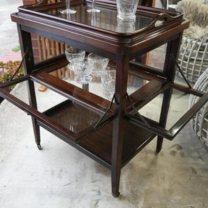 May include: A dark brown wooden serving trolley on wheels featuring a removable top tray with handles, a middle shelf with fold-out glass side panels, and a bottom shelf. The trolley holds a clear glass decanter and several clear glass coupes.