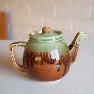 May include: A ceramic teapot featuring a rounded body with a glossy glaze that transitions from a light sage green at the top to a deep, rich brown at the base. The teapot includes a matching lid with a small knob and a curved handle and spout.