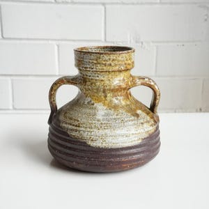 May include: A ceramic vase with two handles, featuring a wide, rounded base with horizontal ridges in a dark brown finish. The upper portion and neck are glazed in a mottled, textured blend of mustard yellow, tan, and cream tones.