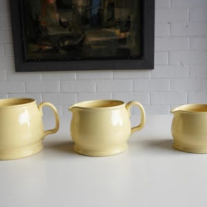 May include: Three pale yellow ceramic jugs of different sizes are displayed. Each jug has a handle and a spout. The background features a framed painting and a white brick wall. Measurements are in cm.