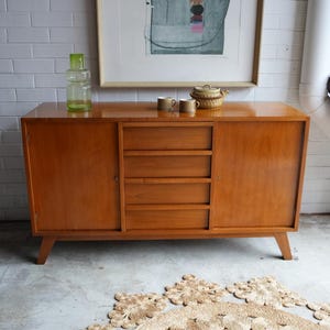 May include: A rectangular wooden sideboard featuring two side cabinets with doors and a central column of four horizontal drawers. The furniture piece has a smooth light brown finish and is supported by four angled wooden legs.